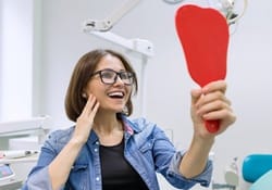 A middle-aged woman wearing glasses looking at her new smile in the mirror after receiving metal-free restorations in Downers Grove